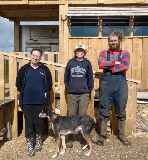 two women, a man and a dog standing outside a wooden building